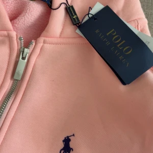 Rosa hoodiejacka från Polo Ralph Lauren - Snygg ljusrosa hoodiejacka från Polo Ralph Lauren med dragkedja, huva med snörning och klassisk broderad logga på bröstet. Jackan har kängurufickor och är tillverkad i mjuk bomull, perfekt för chill dagar.