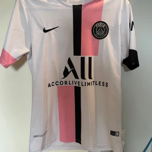 PSG Messi 30 Nike fotbollströja - Paris Saint-Germain fotbollströja med Messi 30-tryck på ryggen. Vit bas med svarta och rosa vertikala ränder, svarta och rosa detaljer på ärmarna. PSG-emblem på bröstet och Nike-logga. Tillverkad i lätt och ventilerande material, perfekt för match eller träning.