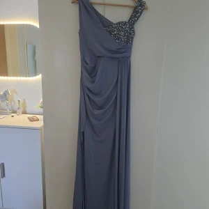 Lång blå one shoulder klänning - Snygg långklänning i blått med one shoulder-design och glittriga paljettdetaljer på axelbandet och bysten. Klänningen har draperad framsida och ett elegant fall. Perfekt för bal eller festliga tillfällen.