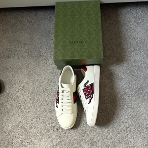 Gucci Ace skor - Gucci Ace sneakers i vitt skinn med ikonisk röd och svart orm brodyr på sidan, grönt och rött band samt metallicröd och metallicgrön häl med Gucci-logga. Klassisk rund tå, snörning och platt sula. Perfekta för dig som vill sticka ut med lyxig streetstyle.