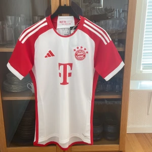 FC Bayern München Adidas matchtröja S 2023-2024 - Säljer en officiell FC Bayern München matchtröja från Adidas i storlek S. Tröjan är vit med röda detaljer, korta ärmar och klubbmärke samt Adidas-logga på bröstet. Tillverkad i lätt och ventilerande funktionsmaterial, perfekt för fotboll eller supporterliv.