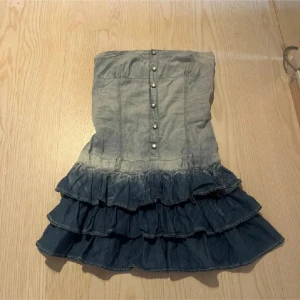 Jeansklänning med volanger och knappar - Supersöt kort jeansklänning i två nyanser av blått med volanger nertill och knappar framtill. Axelbandslös modell med smock i ryggen för skön passform. Perfekt för dig som gillar en cool och lekfull stil. 