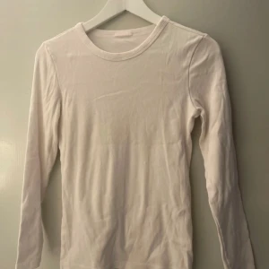 Vit ribbad långärmad tröja - En basic vit långärmad tröja i ribbad bomull. Tröjan har rund halsringning och sitter tajt mot kroppen, perfekt att styla med jeans eller kjol. Enkel och stilren design som funkar året runt. Har en liten fläck som syns på bilden.