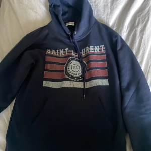 Mörkblå Saint Laurent hoodie - Snygg mörkblå hoodie från Saint Laurent med tryck i rött och vitt framtill. Klassisk huva med snörning och känguruficka. Stort tryck med texten 'Saint Laurent' och grafiska ränder ger en cool vintage vibe. Perfekt för dig som gillar streetstyle.