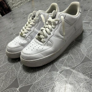 Nike Air Force 1 vita sneakers 46 - Klassiska Nike Air Force 1 sneakers i helvitt skinn med perforerad tå, rund tå och platt sula. Snygg och tidlös modell med snörning och diskret Nike-logga på sidan. Perfekta för dig som gillar en clean och stilren look.
