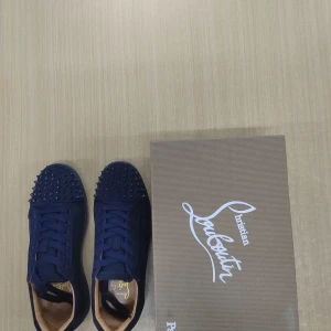 Mörkblå Christian Louboutin sneakers - Snygga mörkblå sneakers från Christian Louboutin i mocka med coola nitar över tån. Skorna har snörning, rund tå och den klassiska röda sulan. Perfekta för dig som vill sticka ut med exklusiv streetstyle.