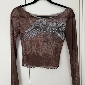 Brun mesh topp med vingar och hjärta - Säljer en brun mesh topp med långa ärmar och croppad passform. Framsidan har ett stort tryck med vingar, hjärta och rosor i grått och vitt. Toppen har vågig kant nertill och är genomskinlig med coola detaljer även på ryggen.