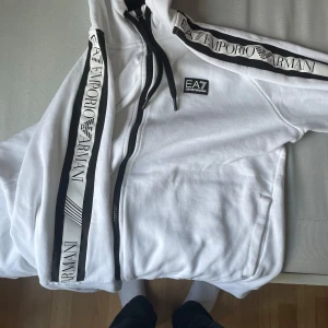 Vit zip-hoodie från Emporio Armani - Snygg vit hoodie från Emporio Armani EA7 med svarta detaljer och dragkedja. Logga på bröstet och breda band med Emporio Armani-tryck längs ärmarna. Mjuk bomullskänsla, huva med snörning och två fickor framtill. Perfekt för en clean och sportig look.