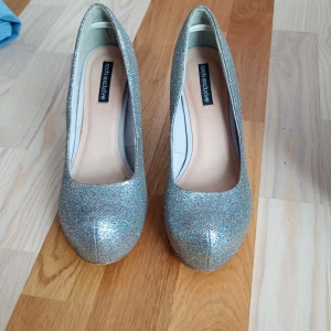 Glittriga pumps från Roots, strl 37 - Snygga pumps från Roots i storlek 37, täckta med glittrigt silverfärgat material. Skorna har rund tå och hög klack, perfekta för att sticka ut. Märke: Roots, köpta på Eurosko. Riktigt statement-piece för dig som gillar glitter.De är perfeckta om du är ett sabrina   fan.Detta märke de här kommer ifrån är jätte exclusive!