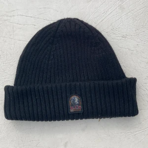 Svart ribbstickad mössa Parajumpers - Svart ribbstickad mössa från Parajumpers med uppvikt kant och klassisk PJS-logga framtill. Tillverkad i mjukt ullmaterial som håller dig varm. Perfekt för kalla dagar och varma dagar med. Överlag väldigt snygg mössa!