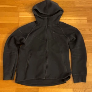 Svart Nike Tech fleece - Svart fleecejacka med dragkedja och huva, perfekt för kyliga dagar. Jackan har två fickor med dragkedja och en enkel, stilren design. Långärmad modell med något längre ryggslut för extra komfort. Passar dig som gillar en clean och sportig look.