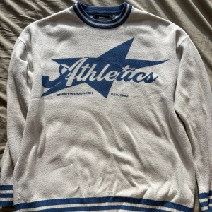 Vit och blå Athletics-tröja Bershka - Säljer en vit långärmad tröja från Bershka med blå detaljer och stor blå stjärna samt texten 'Athletics Brentwood High' framtill. Tröjan har ribbade muddar och krage med blå och vita ränder. Perfekt för en sportig och avslappnad stil.