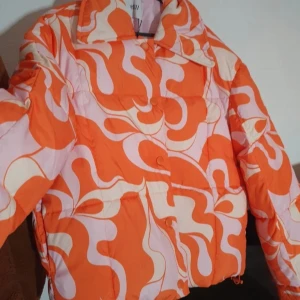 Färgglad pufferjacka från Envii M/L - Säljer en riktigt cool pufferjacka från Envii i storlek M/L. Jackan har ett unikt, vågigt mönster i orange, rosa och beige. Oversized fit med stora krage och tryckknappar framtill. Perfekt statement-piece för dig som vill sticka ut i höst och vinter!