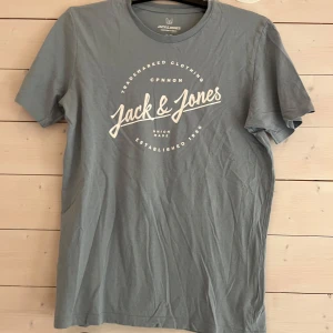 Grå t-shirt från Jack & Jones - Snygg grå t-shirt från Jack & Jones med vit logga och tryck framtill. Klassisk rund halsringning och korta ärmar. Tillverkad i mjuk bomull som känns skön mot huden. Perfekt för en avslappnad och stilren look.