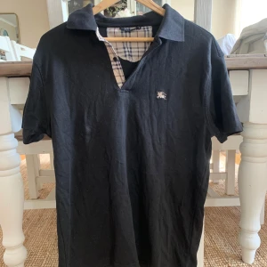 Svart pikétröja från Burberry - Svart pikétröja från Burberry med klassisk krage och korta ärmar. Insidan av kragen har det ikoniska rutiga Burberry-mönstret i beige, blått och rött. Liten broderad Burberry-logga på bröstet. Perfekt för en clean och stilren look.