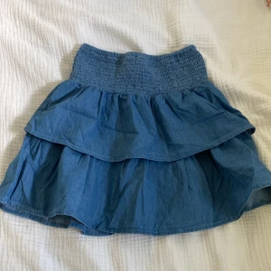 Blå volangkjol i jeans - Supersöt blå kortkjol i jeansmaterial med bred resår i midjan och dubbla volanger. Perfekt för dig som gillar en avslappnad men ändå trendig stil. Lätt att matcha med både sneakers och boots.