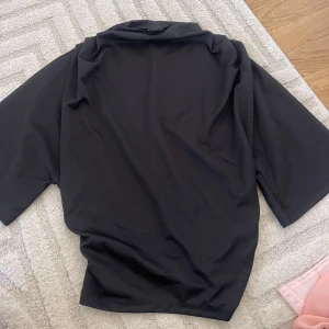 Svart polotröja med kort ärm - En svart polotröja med korta ärmar och avslappnad passform. Perfekt för dig som gillar en clean och stilren look. Materialet känns mjukt och lätt mot huden, och den höga kragen ger en trendig touch till outfiten.