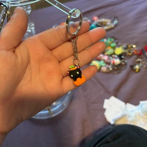 Nyckelring med svart katt och pumpa - Söt nyckelring med en liten svart katt som sitter i en orange pumpa. Katten har vita ögon, rosa öron och ett grönt blad på huvudet. Perfekt accessoar för att piffa upp väskan eller nyckelknippan.
