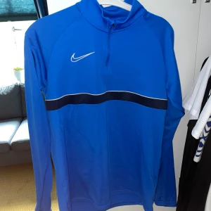 Blå Nike Dri-FIT half zip tröja - Nike Dri-FIT tröja i klarblått med svart och vit rand över bröstet. Långärmad modell med half zip och hög krage. Perfekt för träning eller sportiga dagar. Materialet är lätt och andas, vilket gör den skön att ha på sig under aktivitet.