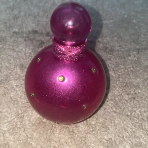 Britney Spears Fantasy parfym 100ml - Fantasy av Britney Spears är en ikonisk parfym i en rund, glittrig rosa flaska med gröna stenar och dekorativ hals. Flaskan rymmer 100 ml och har en lyxig, ungdomlig design som verkligen sticker ut på hyllan. Perfekt för dig som gillar söta och fruktiga dofter. De är ca hälften kvar. Köpte den för nästan 300 kr 