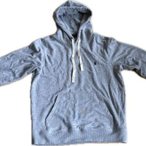 Grå hoodie från Polo Ralph Lauren - Klassisk grå hoodie från Polo Ralph Lauren med vit snörning och broderad logga på bröstet. Tröjan har känguruficka, huva och är tillverkad i mjuk bomullsmix. Perfekt för en chill och stilren look.