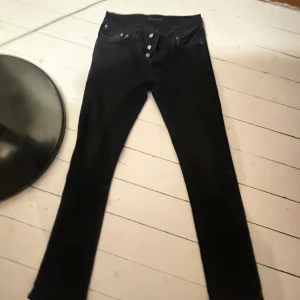 Svarta jens från Nudie Jeans - Svarta  jeans från Nudie Jeans med klassisk femficksdesign och knappgylf. Jeansen är i stretchig bomull och har en tight passform som sitter snyggt längs benen. Perfekta för dig som gillar en stilren och enkel look.