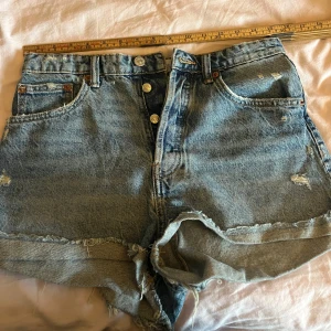 Jeans shorts zara - Snygga  jeansshorts i ljusblå denim med slitna detaljer och råa kanter. Klassisk femficksmodell med knappar framtill och uppvikta benslut. Perfekta för varma dagar och en avslappnad stil.