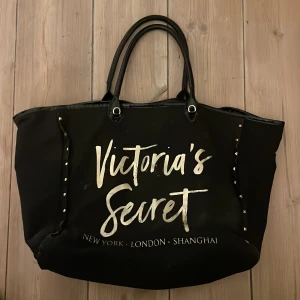 Stor svart shoppingväska Victoria's Secret - Snygg och rymlig svart shoppingväska från Victoria's Secret med stora handtag och coolt tryck i vitt och guld på framsidan. Väskan har nitar på sidorna och innerfack med dragkedja. Perfekt för dig som vill ha plats för allt och lite till. Fläck på baksidan (inte testat att tvätta bort) men syns inte när man har på sig den.