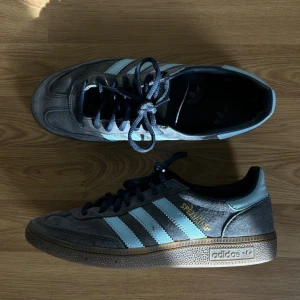 Adidas Spezial sneakers grå/blå mocka - Spliter nya Adidas Spezial sneakers i grå mocka med ljusblå detaljer och klassiska tre ränder på sidorna. Skorna har mörkblå snörning, gummisula och guldfärgad Spezial-logga. Perfekt för dig som gillar retrostil och streetwear.