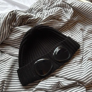 Svart Goggle-mössa från C.P. Company - Svart ribbstickad mössa från C.P. Company med ikoniska svarta goggle-linser framtill. Mössan är i tjockt, stickat material och har en bred uppvikt kant. Perfekt för dig som vill sticka ut med streetwear-vibe.
