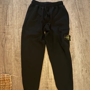 Svarta mjukisbyxor från Stone Island - Svarta mjukisbyxor från Stone Island med klassisk logga på benet. Byxorna har snörning i midjan och en praktisk cargoficka på sidan. Perfekta för en chill och avslappnad stil. Tillverkade i mjukt bomullsmaterial.