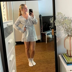 Blommig omlottklänning/playsuit från Chiquelle - Omlottklänning/playsuit från Chiquelle i ljusblått med små vita och gula blommor. Klänningen har långa ärmar med volangdetalj vid ärmslut, knyt i midjan och v-ringning. Är en playsuit som ser ut aom en klänning framifrån, hekt oanvänd.