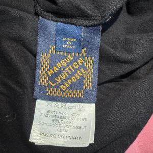 Svart Louis Vuitton t-shirt med röd logga - Svart t-shirt från Louis Vuitton med klassisk rund hals och korta ärmar. På bröstet finns en broderad röd LV-logga som ger en clean och exklusiv vibe. Tillverkad i mjuk bomull och made in Italy – perfekt för dig som gillar stilrena designer med lyxig touch.