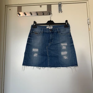 Blå jeanskjol med slitningar - Snygg blå jeanskjol med slitna detaljer och fransig nederkant. Klassisk femficksmodell med dragkedja och knapp framtill. Perfekt för en avslappnad och trendig look. Från märket Odd Molly i storlek 1 som ungefär motsvarar en Storlek S. Mycket fint skick, jag hittar inga synlig anmärkningar eller defekter. 