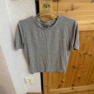 Grå ribbad topp med pärlor  - Grå ribbad topp från Indiska med korta ärmar och rund hals. Toppen är dekorerad med små pärlor framtill som ger en extra touch. Perfekt att styla med jeans eller kjol för en trendig look.