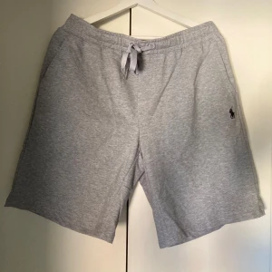 Grå shorts från Polo Ralph Lauren - Nya, aldrig använda ljusgrå shorts från Polo Ralph Lauren med klassisk broderad logga på benet. Mjukt bomullsmaterial 97%, elastisk midja med snörning och fickor på sidorna samt en bakficka.                                                        Storlek L. (M)                                                          längd hela shortsen 49 cm,  bredd resåren ca 41cm eleastrisk till 54 cm diameter på mig är 105cm så borde passa perfekt om man har 90-115 cm runt midjan utan att använda snörning