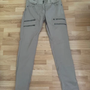 Beiga cargopants från House of Soul - Snygga beiga cargopants från House of Soul med fyra dragkedjor framtill som cool detalj. Byxorna har normal passform, raka ben och klassiska fickor både fram och bak. Perfekta för dig som gillar en stilren men ändå edgy look. Midjan 80-85cm.
