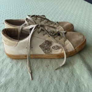 Glittriga sneakers Le Temps des Cerises - Snygga beiga sneakers från Le Temps des Cerises med glittrigt hjärtdetalj på sidan och glittrande snören. Skorna har beige detaljer och en platt sula i gummi. Perfekta för dig som gillar att sticka ut med lite extra glitter i vardagen. De är lite smutsiga men jag ska tvätta dem! 
