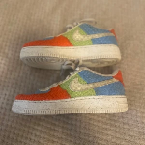 Nike Air Force 1 Hemp i färgblock - Unika Nike Air Force 1 Hemp sneakers med färgblock i blått, grönt, rött och beige. Skorna har en vävd struktur och klassisk vit sula. Swoosh-loggan och snörningen är i beige. Perfekta för dig som vill sticka ut med färg och textur.