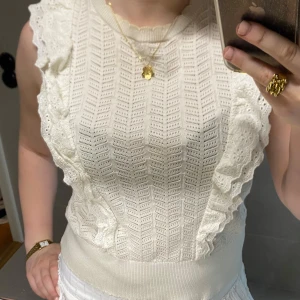 Vit spetsig blus - Supersöt vit blus från Holly & Whyte med broderade volangdetaljer längs axlarna och ett stickat, hålmönstrat tyg. Ärmlös modell med ribbad midja och romantisk känsla. Perfekt för dig som gillar en feminin och trendig stil.