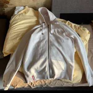 Vit hoodie från A.C.E med dragkedja - Vit hoodie från A.C.E med broderad logga på bröstet och coolt tygmärke på ärmen med texten 'ARE YOU'. Tröjan har hel dragkedja, huva med snörning och ribbade muddar. Snygga detaljer i rött, vitt och blått på snörena och nederkanten. Perfekt för en chill och stilren look. Jag är lite osäker på storleken på tröjan men den är en S eller M men den passar som både och.