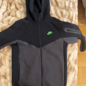 Svart Nike vindjacka med huva XS - Nike vindjacka i svart och mörkgrått med grön logga på bröstet. Jackan har hel dragkedja, huva och en praktisk ficka med dragkedja på ärmen. Perfekt för dig som gillar sportig och clean stil.