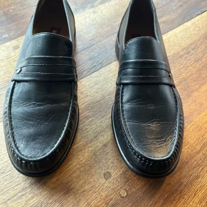 Svarta loafers i skinn från Marco Bossi - Klassiska svarta loafers från Marco Bossi i äkta skinn med extra bred passform. Snygg detalj med liten metall-logga på sidan och diskret sömnad runt tån. Perfekta för dig som gillar stilrena och tidlösa skor med platt sula och rund tå.