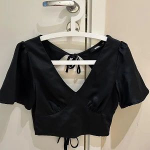 Ny Svart topp från Bik Bok - Snygg svart topp från Bik Bok med korta ärmar och croppad passform. Toppen har v-ringning, knytband i ryggen och en lätt glansig yta. Perfekt för att lyfta din outfit med en trendig touch.
