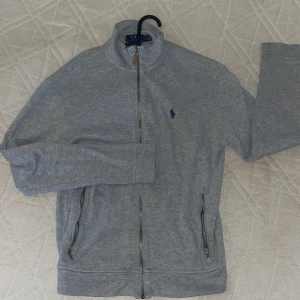 Grå kofta från Polo Ralph Lauren - Snygg grå kofta från Polo Ralph Lauren med dragkedja hela vägen och två fickor med dragkedja. Klassisk broderad logga i mörkblått på bröstet. Mjuk bomullskvalitet och hög krage som ger en clean look. Perfekt till jeans eller joggers.