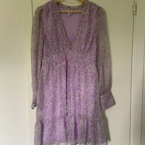 Lila klänning Vero Moda - Supersöt lila kortklänning från Vero Moda med blommigt mönster, v-ringning och långa ärmar. Klänningen har volang nertill samt en dragkedja i sidan. Den är aningen liten i storleken. Höjd: 90cm, Midja: 37cm tvärs över