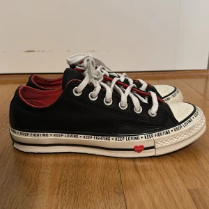 Converse sneakers med hjärtdetaljer - Svarta Converse sneakers i läder med vita snören och röd insida. Texten 'KEEP LOVING KEEP FIGHTING' runt sulan och röda hjärtan på yttersulan och sidan. Klassisk rund tå och platt sula. Perfekt för dig som vill sticka ut med coola detaljer. Storlek 37, skorna är använda men noga rengjorda och inga större defekter, bara små skrapor längst fram på skon.