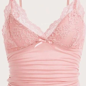 Rosa spetslinnetopp med rosett - Supersöt ljusrosa linnetopp med tunna axelband och v-ringning. Toppen har spetsdetaljer över bysten, en liten rosett framtill och är figurnära med lätt rynk i tyget. Perfekt för dig som gillar romantisk och feminin stil.