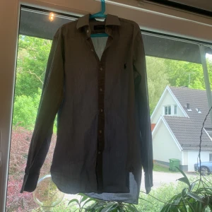Grå skjorta från Polo Ralph Lauren - Snygg grå skjorta från Polo Ralph Lauren med klassisk krage och knäppning framtill. Skjortan har lång ärm och den ikoniska broderade loggan på bröstet. Perfekt för dig som gillar stilrena och tidlösa plagg.