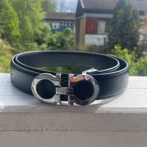 Svart Ferragamo skärp med silverdetaljer - Snyggt svart skärp från Ferragamo i äkta skinn med exklusivt silverfärgat spänne i ikonisk design. Bältet har en stilren och lyxig känsla med präglad yta och tydlig Ferragamo-logga på spännet. Perfekt accessoar för att lyfta din outfit.